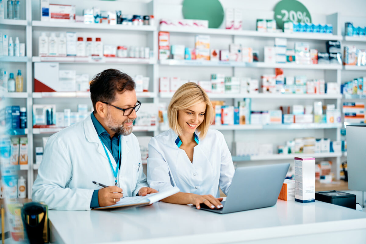 Female pharmacist using laptop while her colleagues is taking notes at the pharmacy counter.